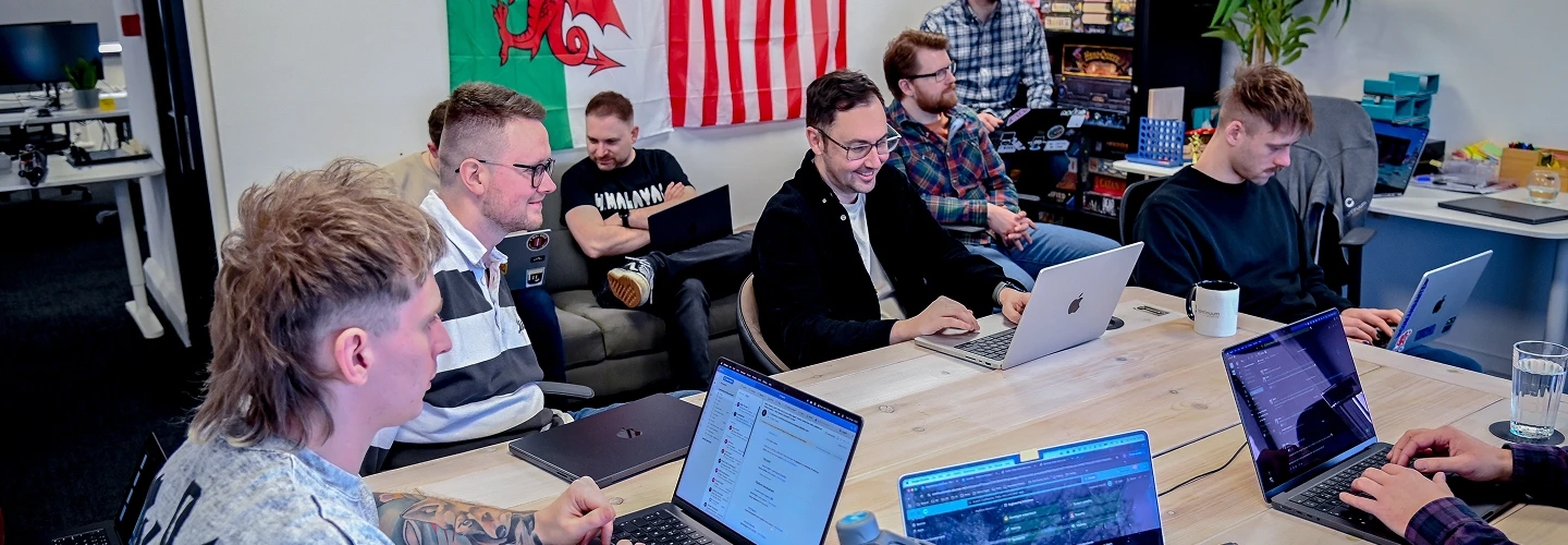 Group of young adults working on laptops around a table in a casual office space with flags on the wall.