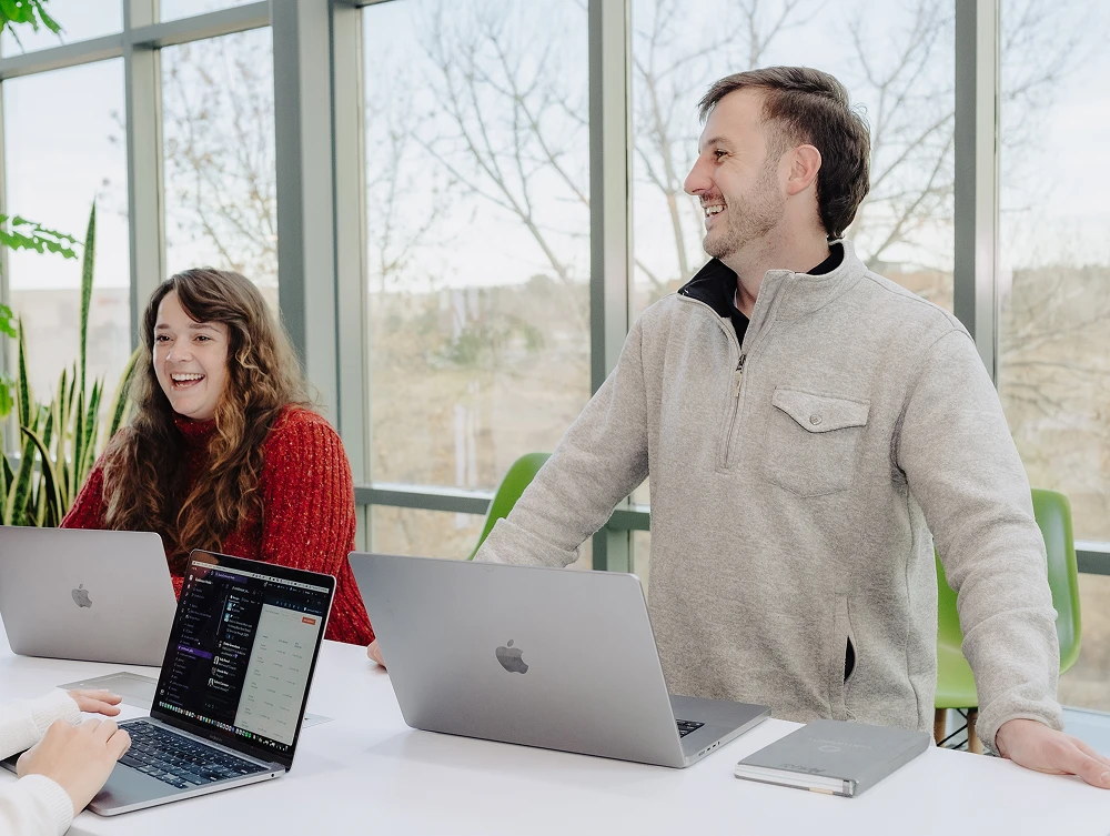 Two people laugh while working on laptops at a table in a bright room with large windows.