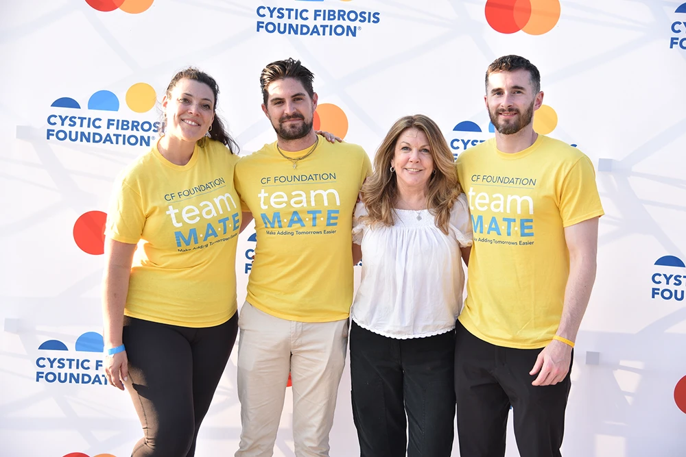 Four people smiling in front of a Cystic Fibrosis Foundation backdrop, three wearing yellow CF Foundation shirts.