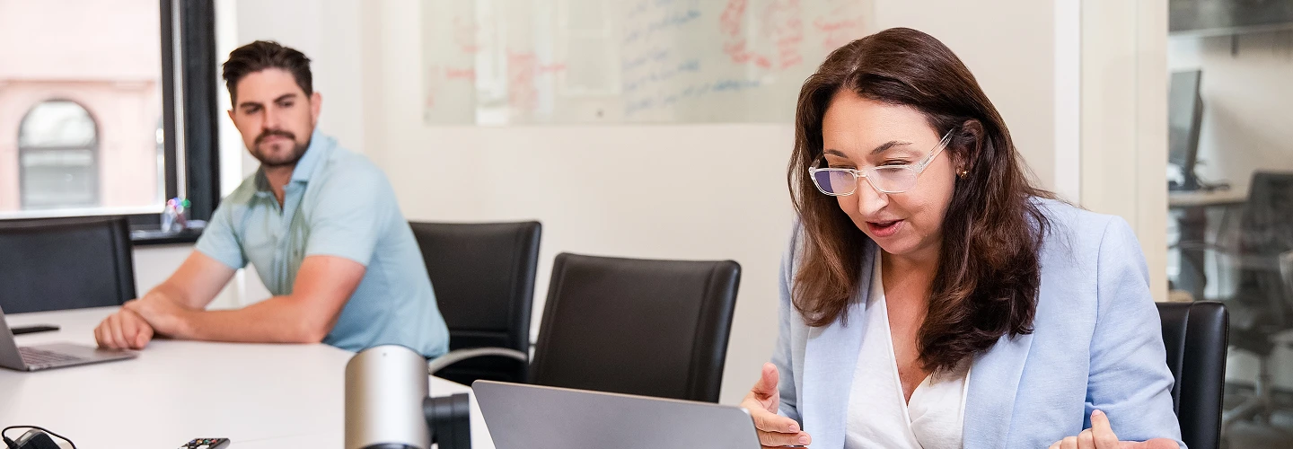 Two colleagues in a meeting room focused on laptop screens with a whiteboard in the background.