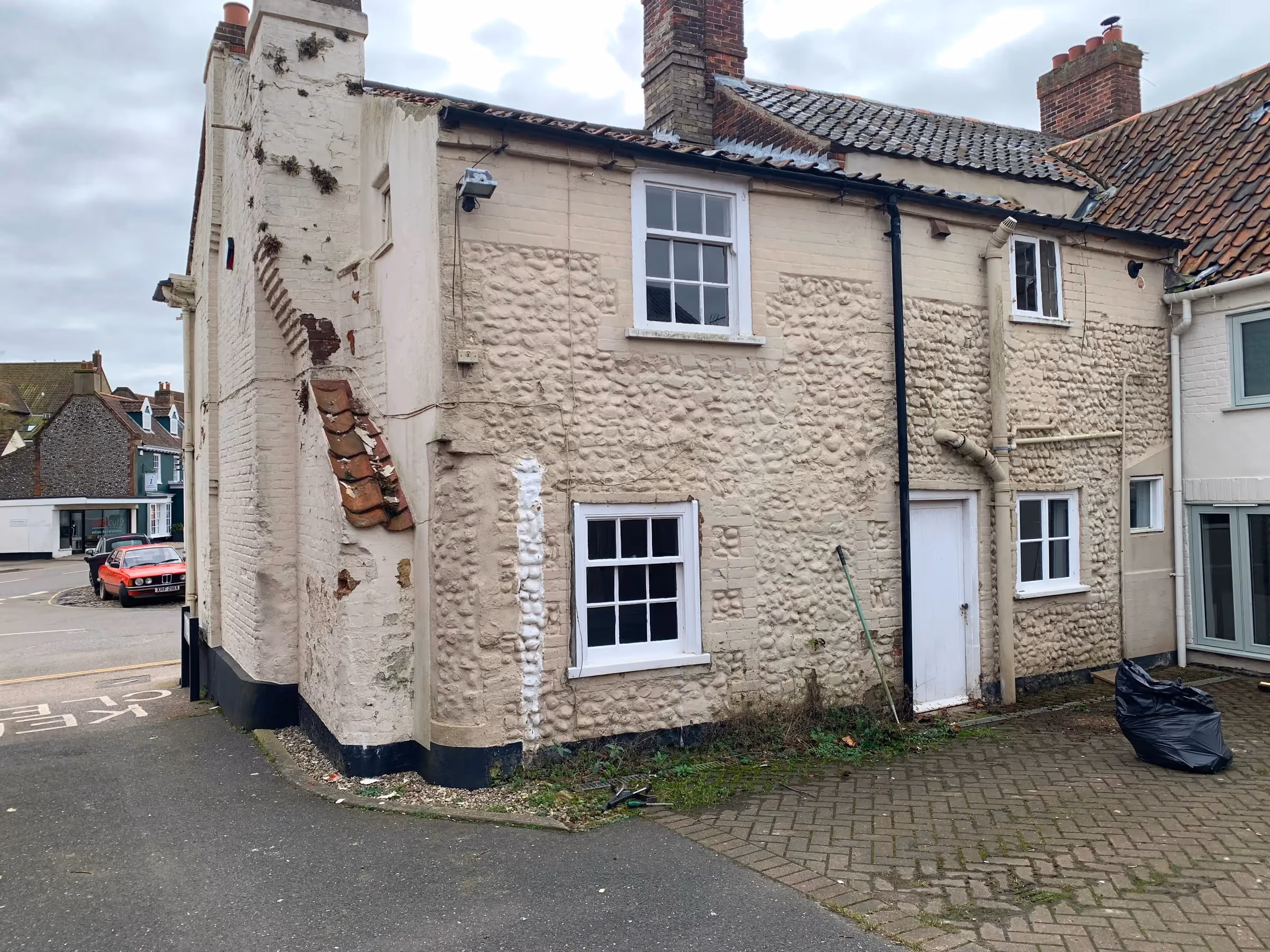 Rear elevation before work, with painted brick and flint walls and weeds growing along the base