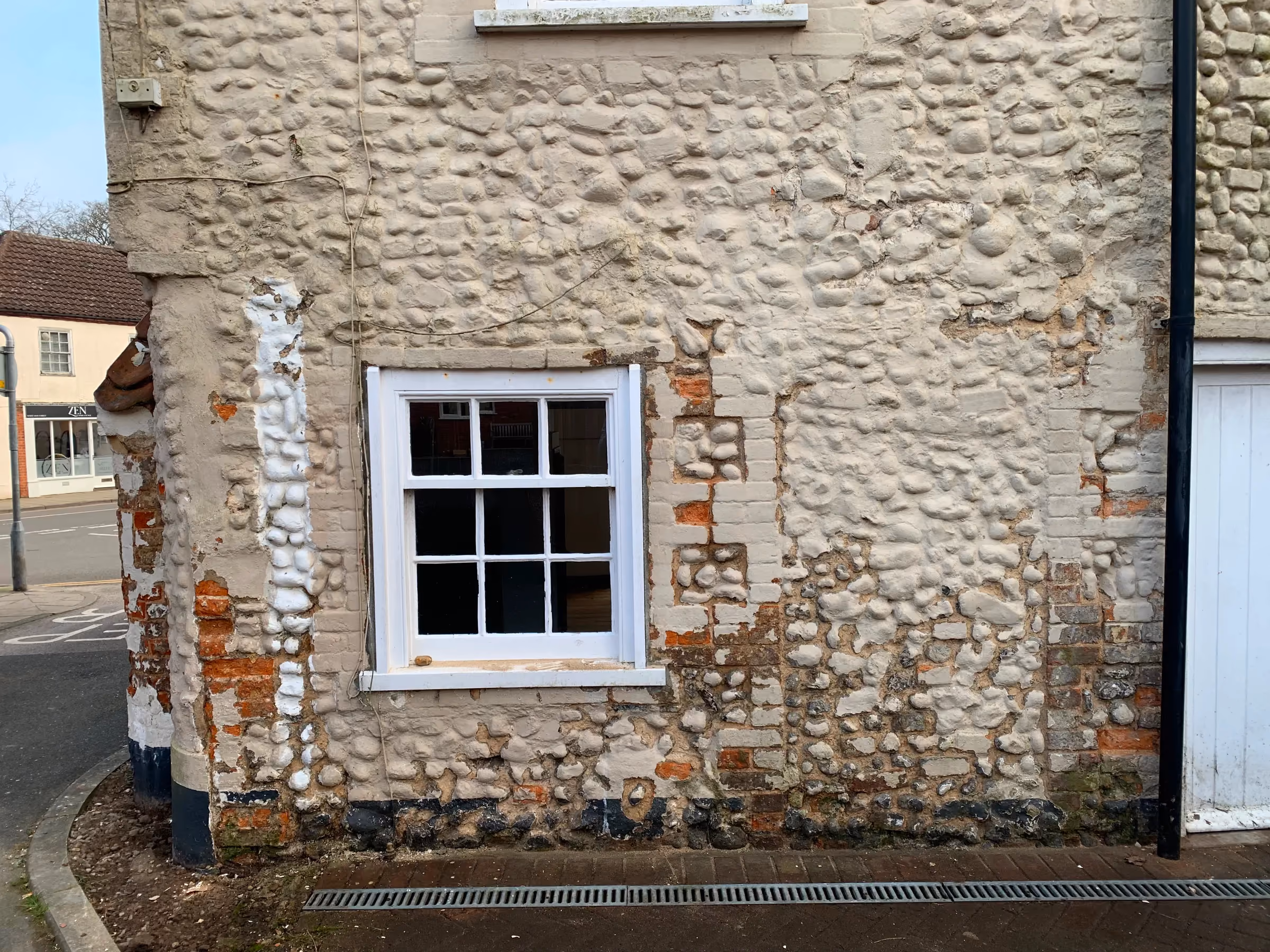 Cleared drain channel running below a sash window, with paint partly stripped from the brick and flint