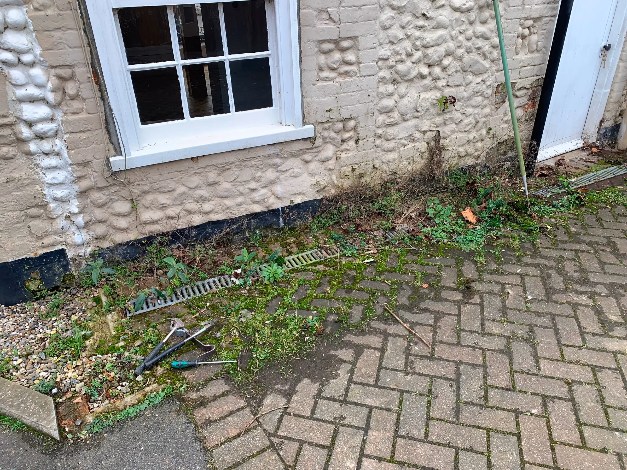 Weeds and soil choking the drain channel along the base of the painted flint wall