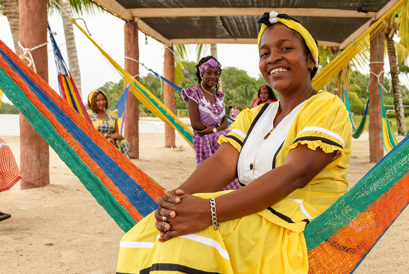 Smiling woman in yellow traditional dress sitting on a colorful hammock under a beach pavilion with other women in traditional clothing in the background.