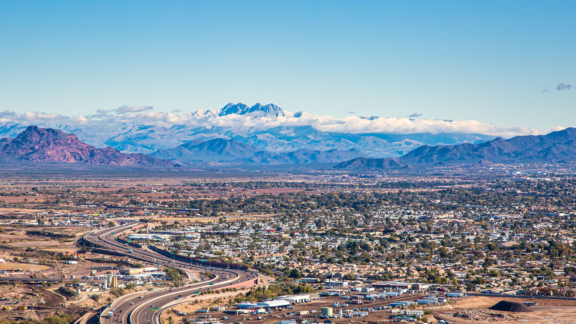 Aerial view of a city with a highway, scattered buildings, and distant mountains under a clear sky.