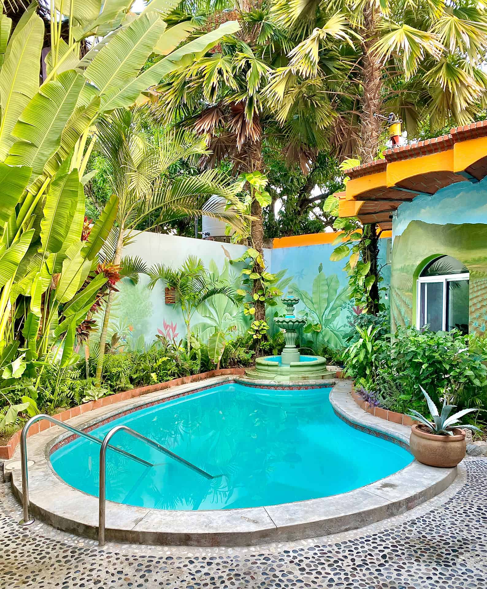 Swimming pool has stairs with handles for easy access. There is a fountain on the far side.