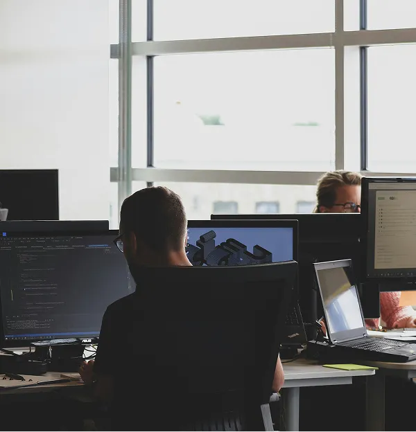 Two individuals work at desks with computers in a bright office space, focusing on their screens. Natural light filters through large windows.