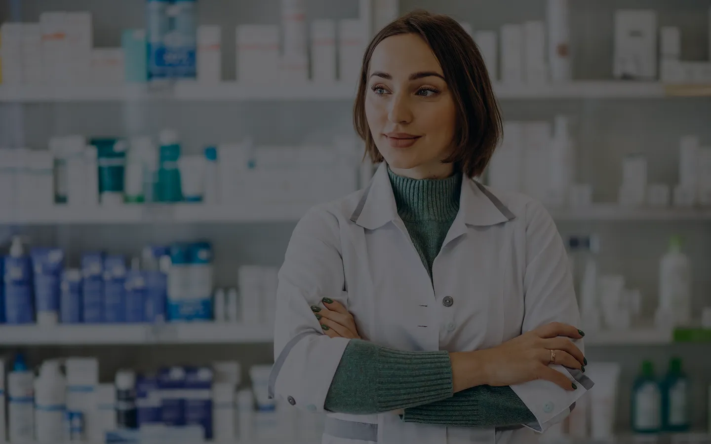 A woman in a white lab coat stands confidently with crossed arms in a pharmacy, shelves of products behind her. She appears professional and content.
