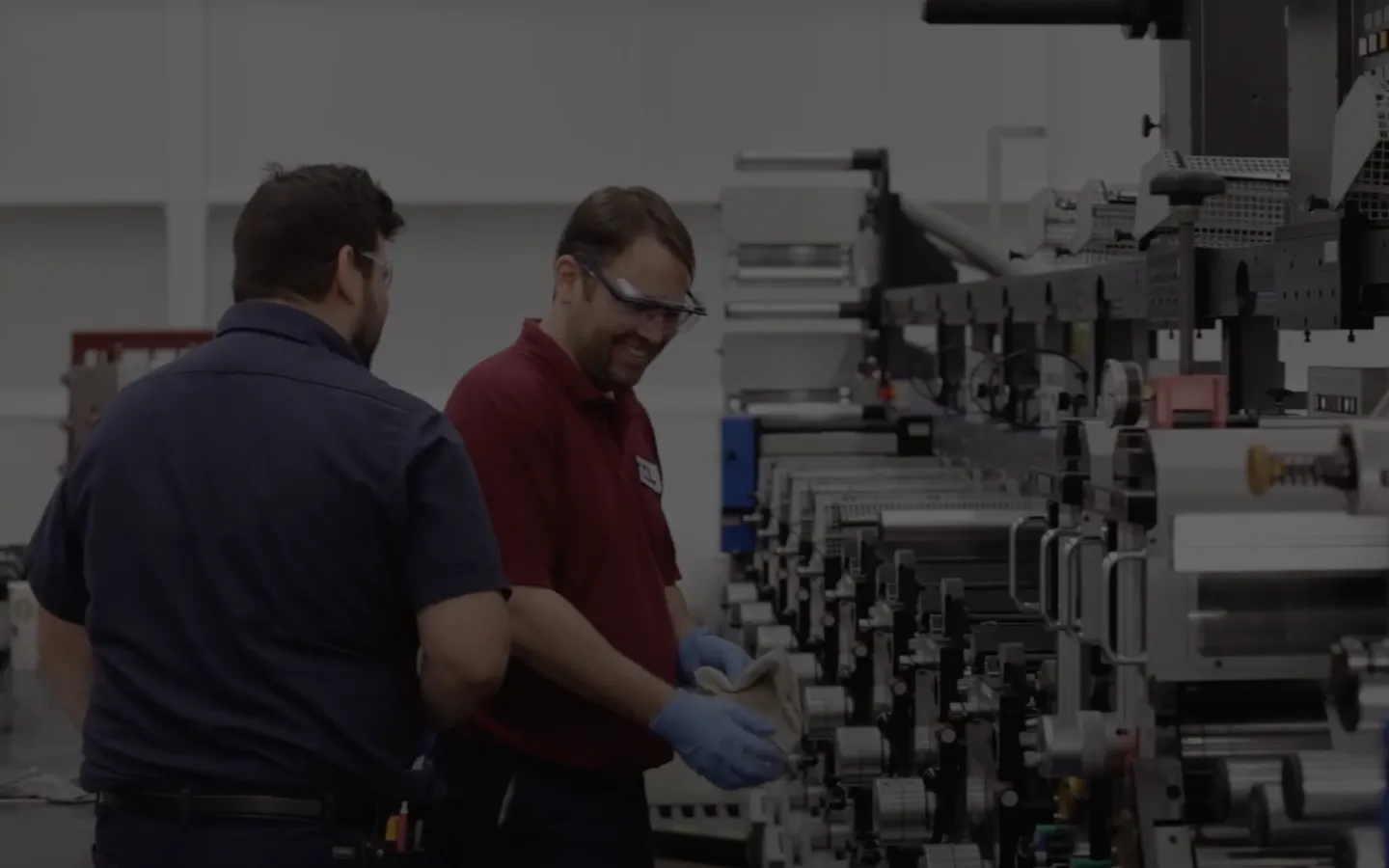 Two men stand next to a large industrial machine in the Printing and Packaging industry; one wears safety glasses and gloves, holding a tool while working on the equipment.
