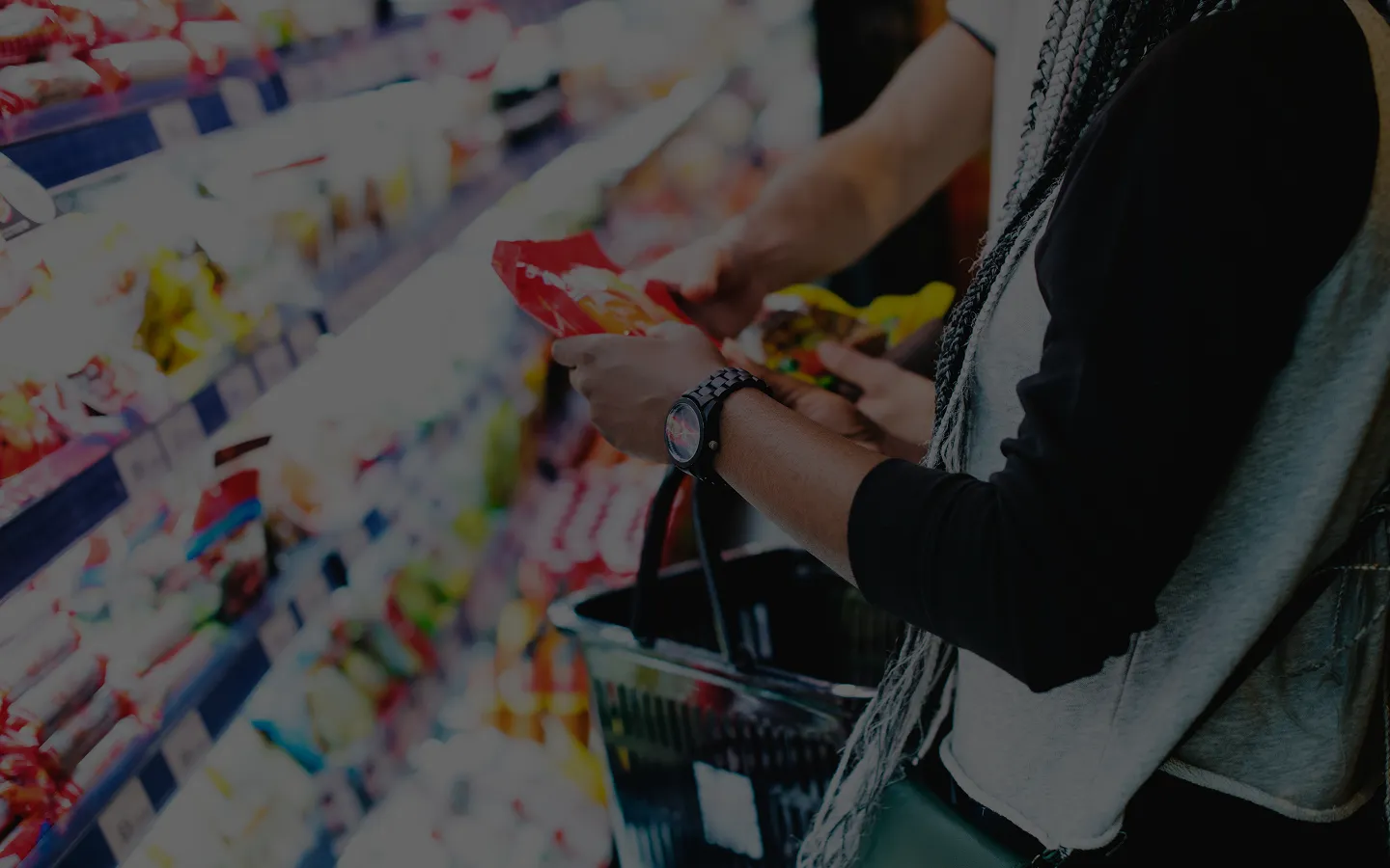 Two people shopping in a grocery store aisle, holding snack packages and a shopping basket—a classic scene from the CPG industry. Shelves stocked with various products are visible in the background.