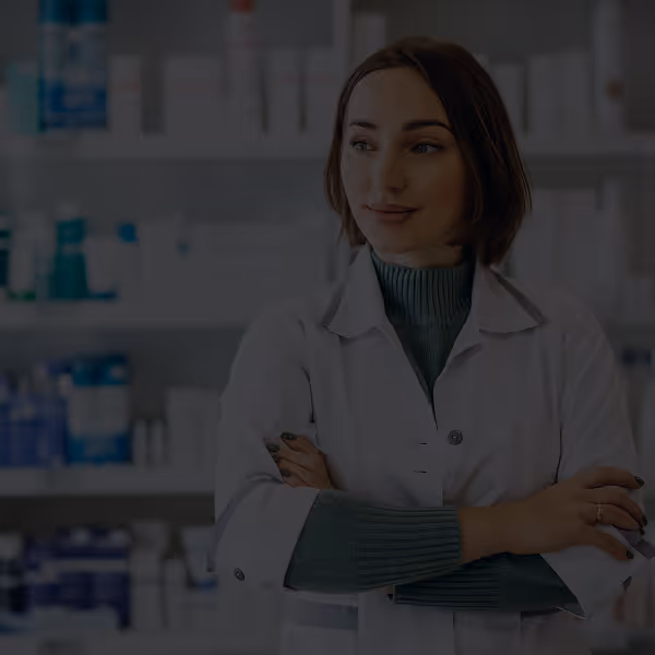 A woman in a white lab coat stands confidently with crossed arms in a pharmacy, shelves of products behind her. She appears professional and content.