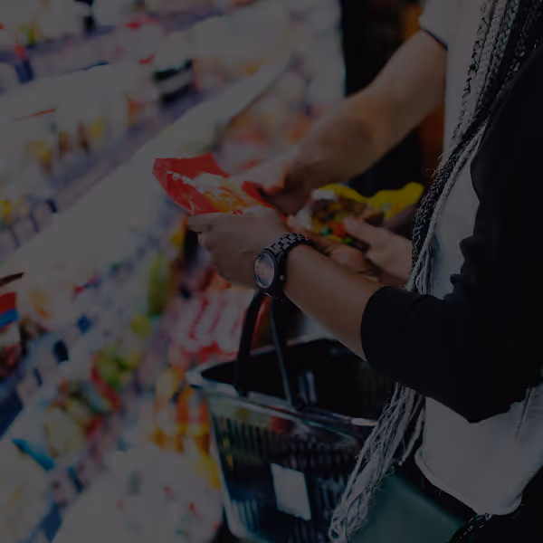 Two people shopping in a grocery store aisle, holding snack packages and a shopping basket—a classic scene from the CPG industry. Shelves stocked with various products are visible in the background.