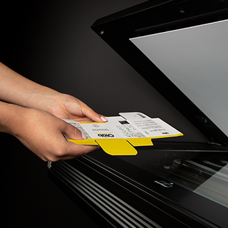 Hands holding a yellow and white print carton being placed into a flatbed scanner on a black surface.