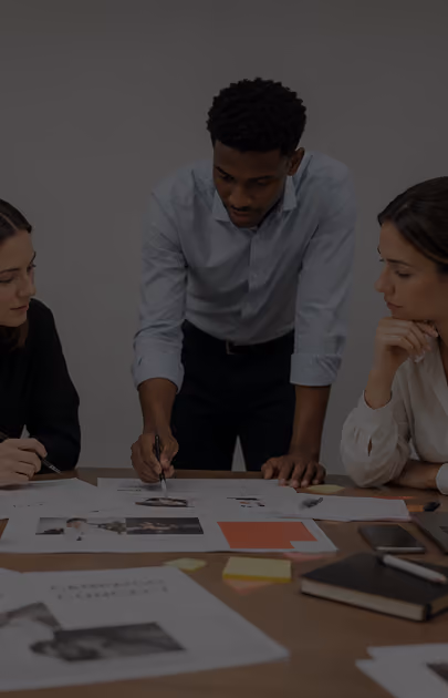 Three people collaborating over documents and photos spread on a table in a meeting.