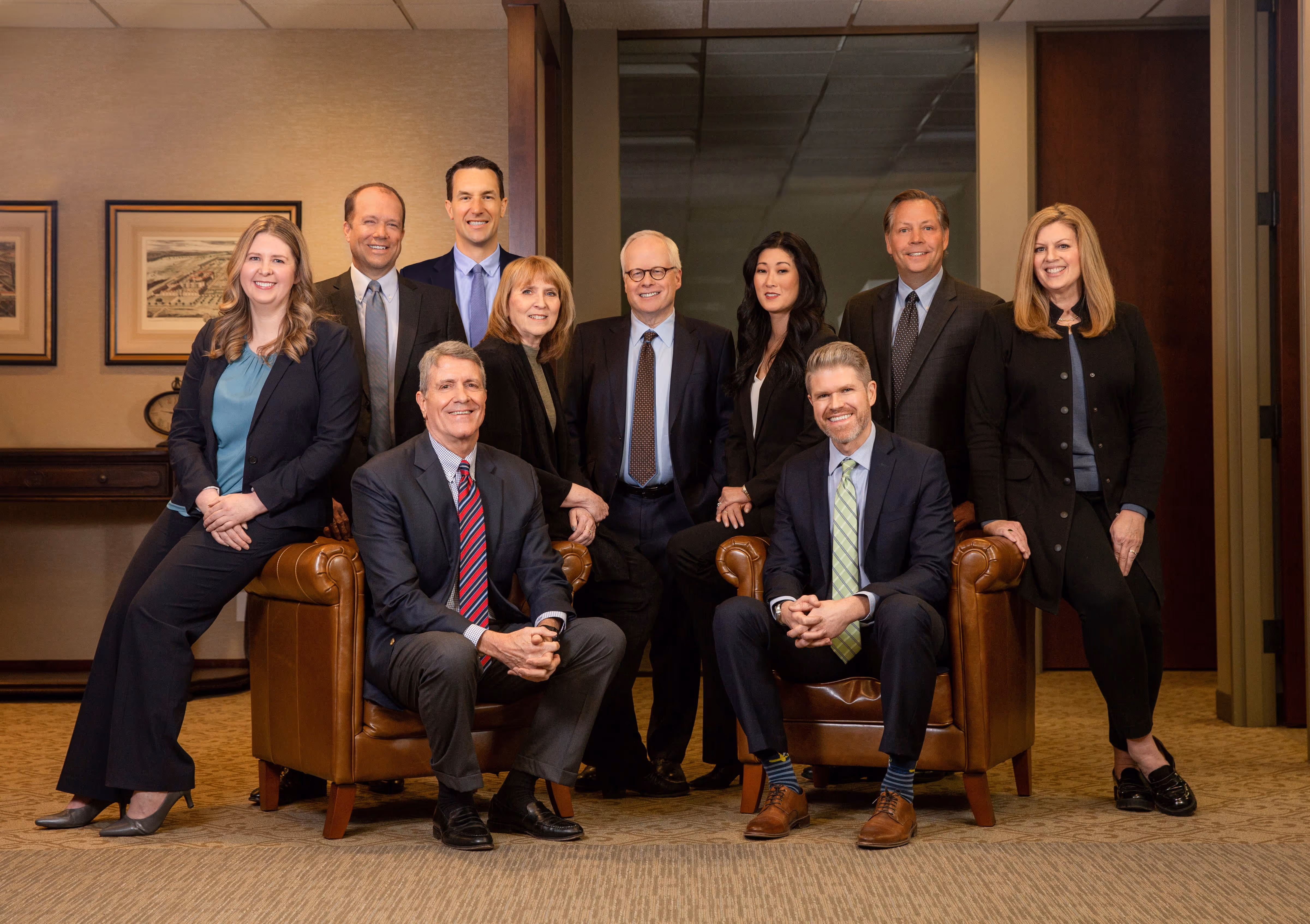 Professional group of ten people in business attire posing indoors with two sitting on leather armchairs and others standing behind.
