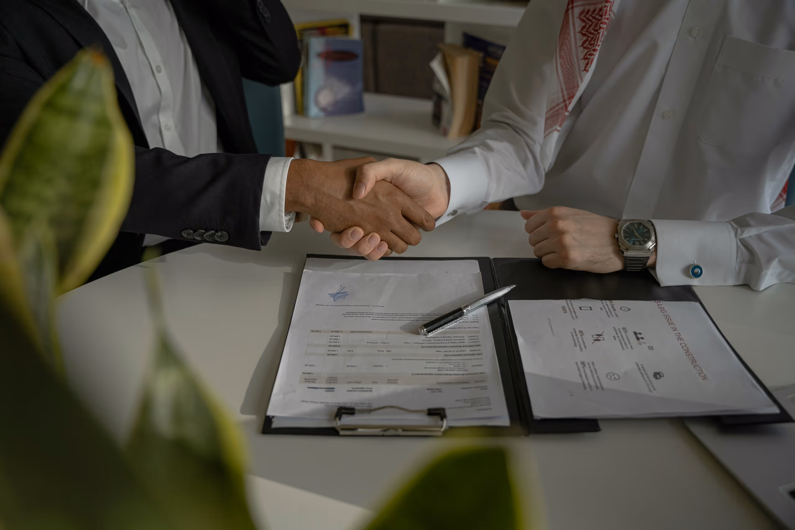 Two men in business attire shaking hands over a desk with documents and a pen.