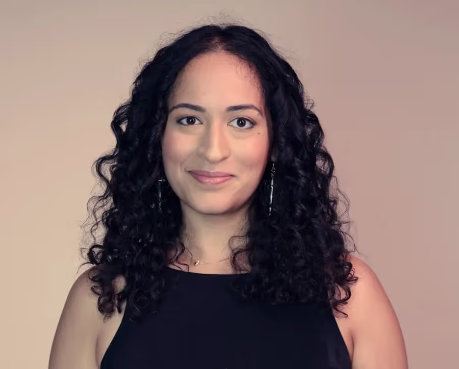 Woman with curly black hair wearing a sleeveless black top, smiling against a beige background.