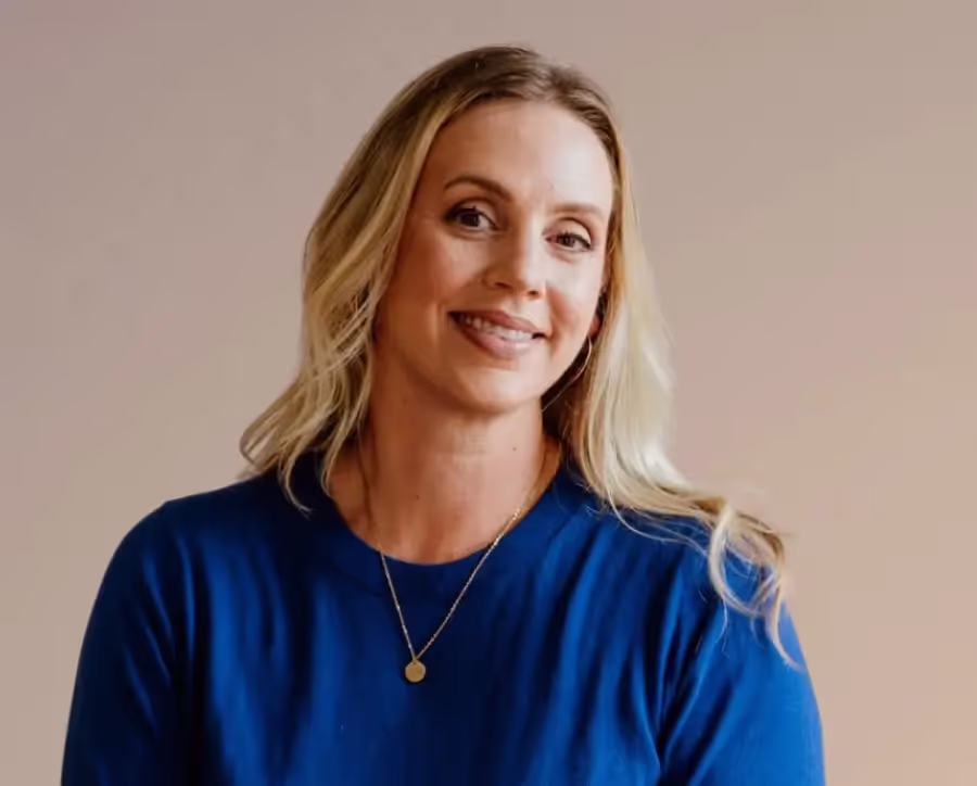 Smiling woman with long blonde hair wearing a blue top and a gold necklace against a beige background.