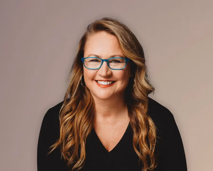 Smiling woman with long wavy hair, teal glasses, and hoop earrings wearing a black top against a neutral background.