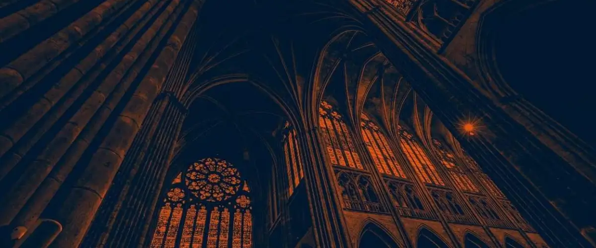 Interior shot of a dark Gothic cathedral with high pillars and stained glass arched windows.