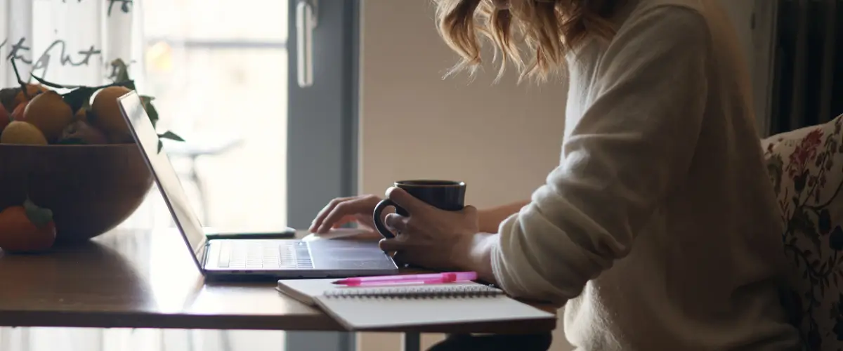 A writer works on their laptop, a mug in one hand, sitting at their kitchen table.