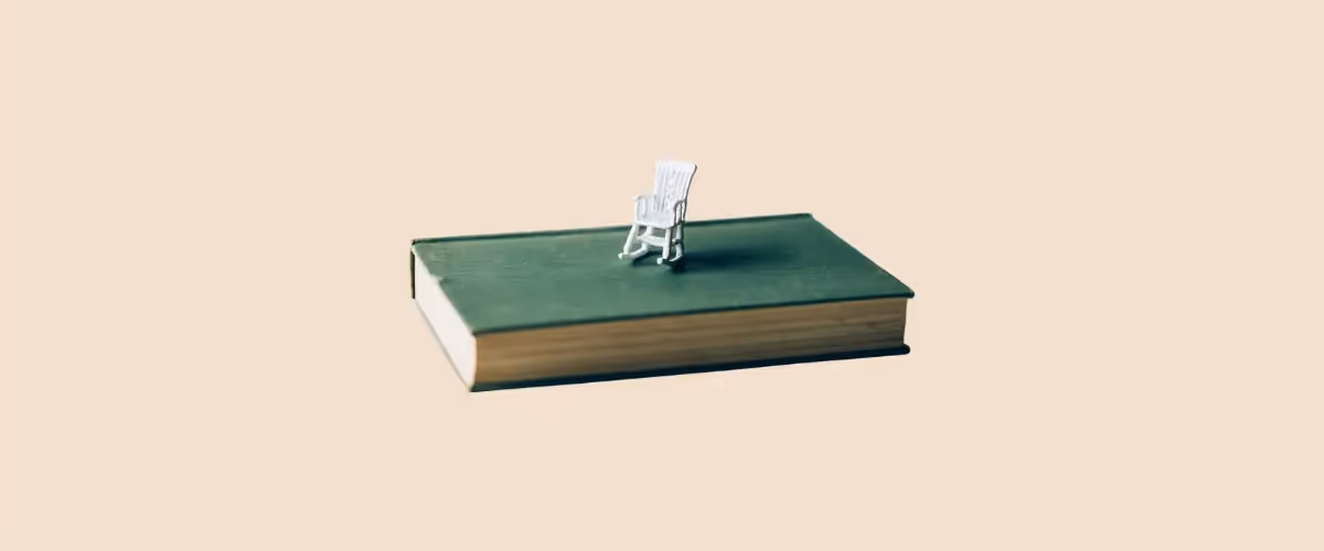 A tiny dolls-house rocking chair in white sits on top of an old hardback book with a green cover.