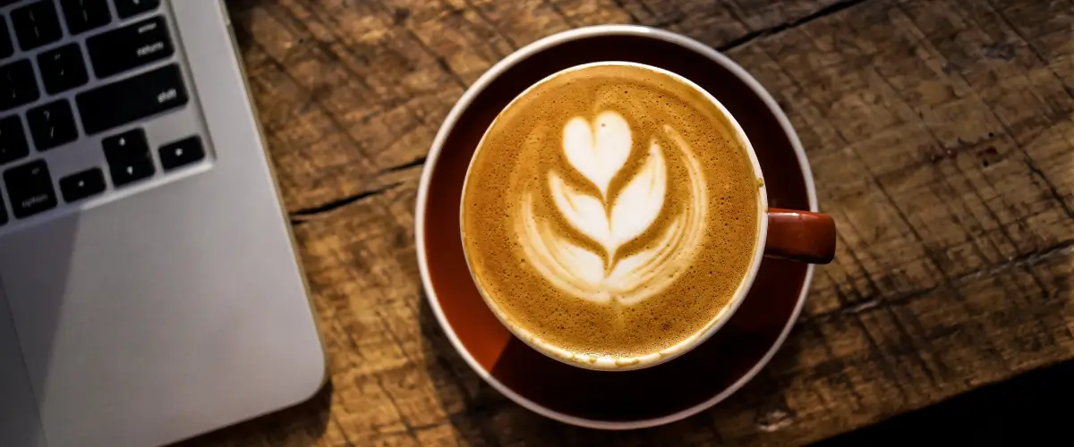 A cup of coffee with a heart drawn in the foam sits on a table beside an open laptop.