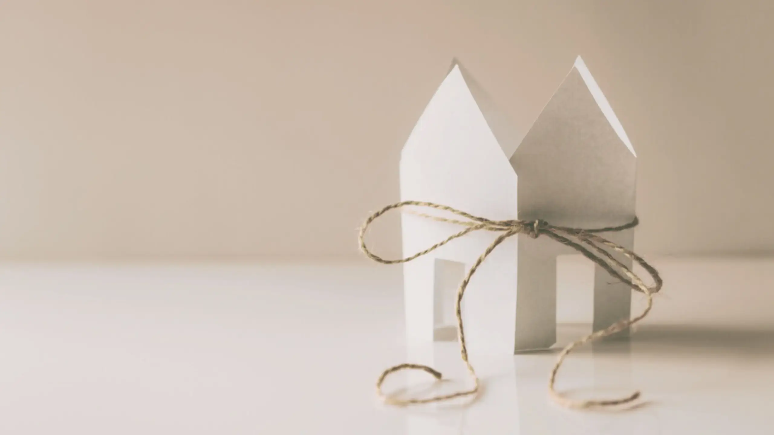 A small house made of folded paper sits on a white surface, held together by a piece of brown string tied in a bow around its middle.