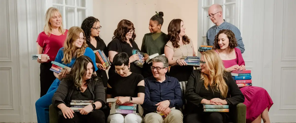 12 of the writing coaches from The Novelry sit and stand in a group, discussing their books, which they are each holding.