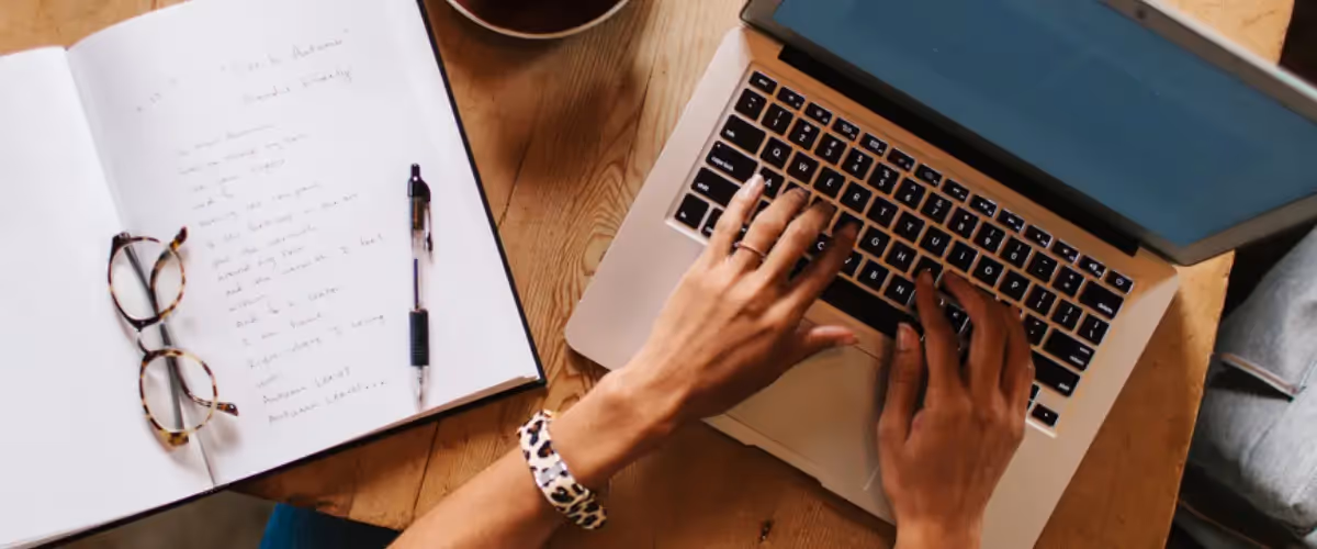 Overhead view of hands typing on a laptop, an open notebook with handwriting in lying beside it.