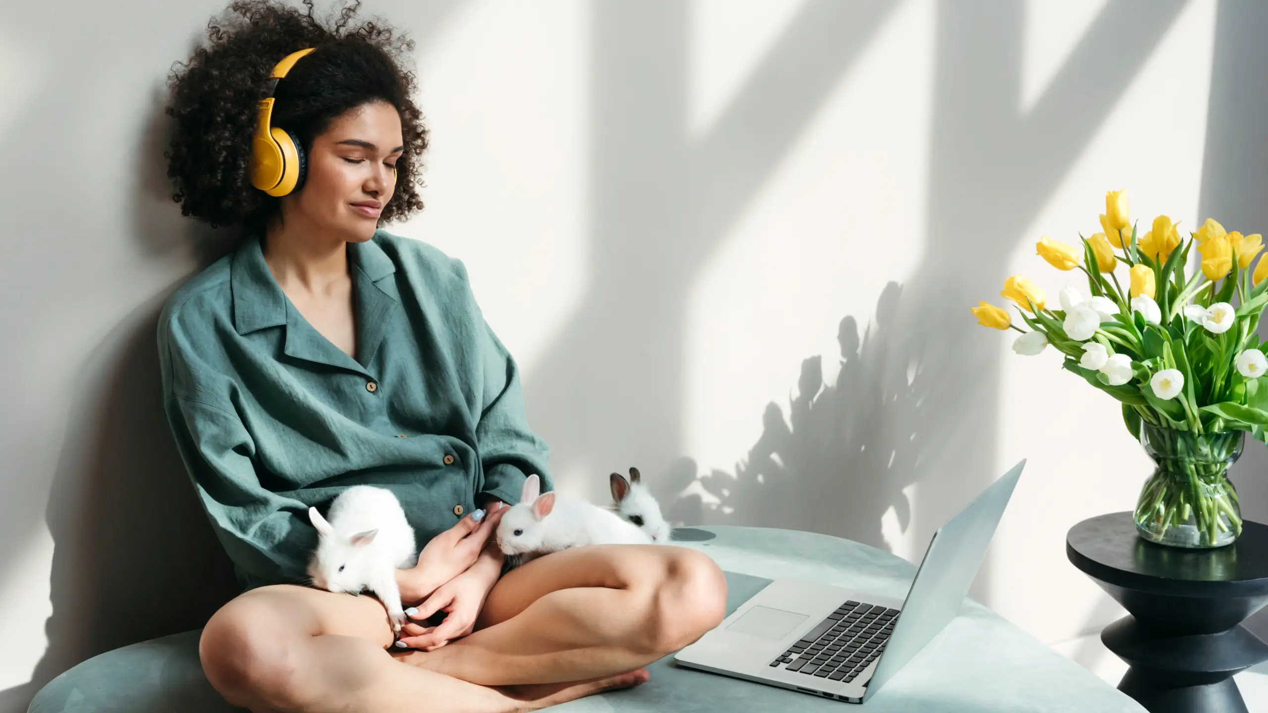 A woman sits in a sun-filled room with headphones on listening to a podcast, with three baby bunnies on her lap.