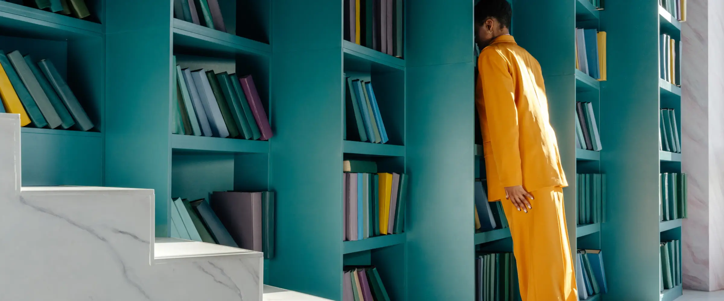 A woman in a yellow suit leans against a row of turquoise bookshelves.