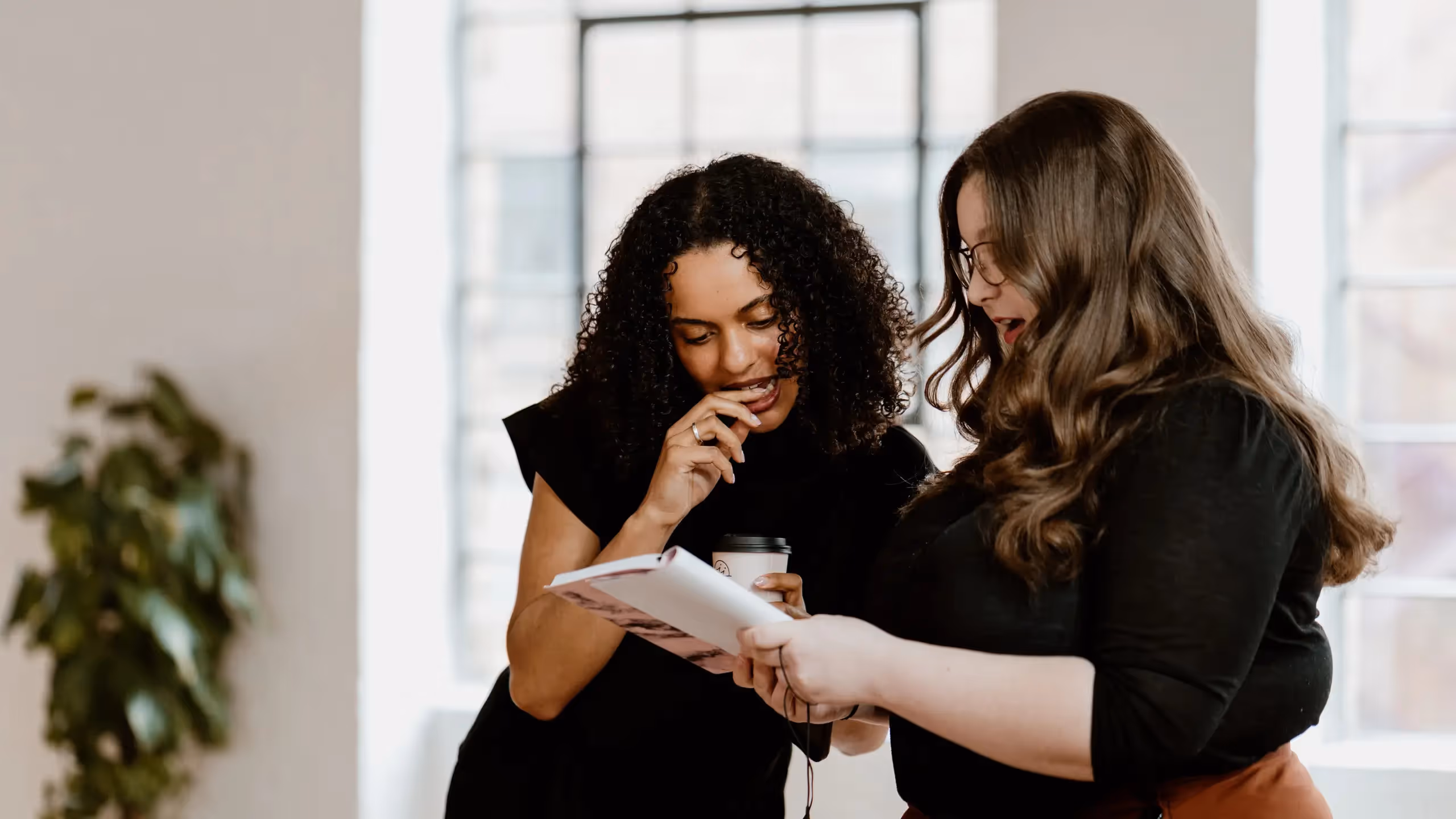 The Novelry editors Sadé Omeje and Georgia Summers discuss a printed manuscript in detail against a large window with a plant beside it.