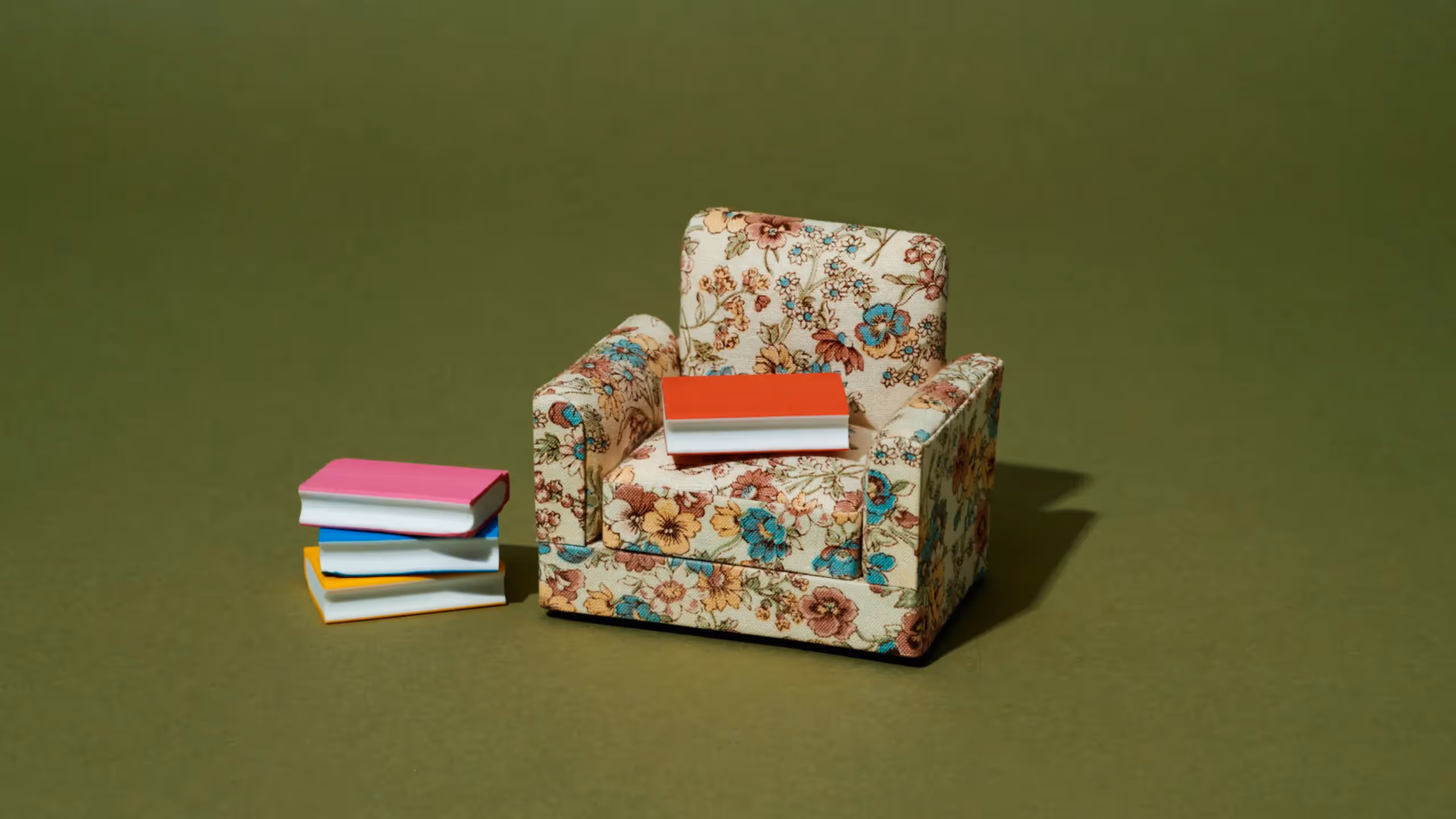 A book lies on a floral armchair against a dark green background, with another small pile of books on the floor beside it.