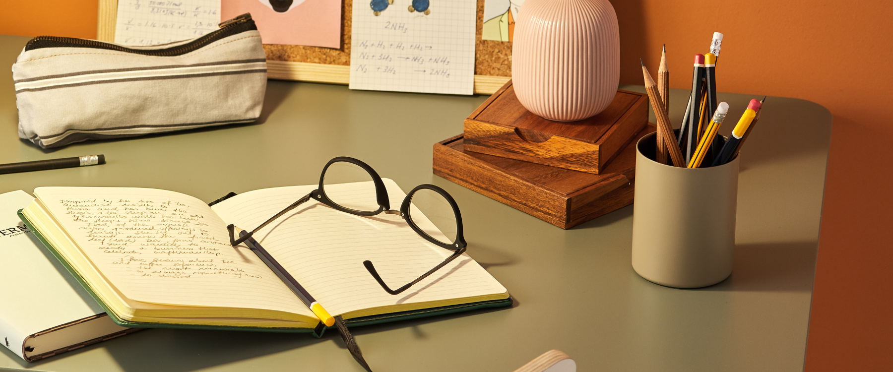 A sage green desk on which stands a pen pot, ornaments, notebook, pen, glasses, and a pencil case.