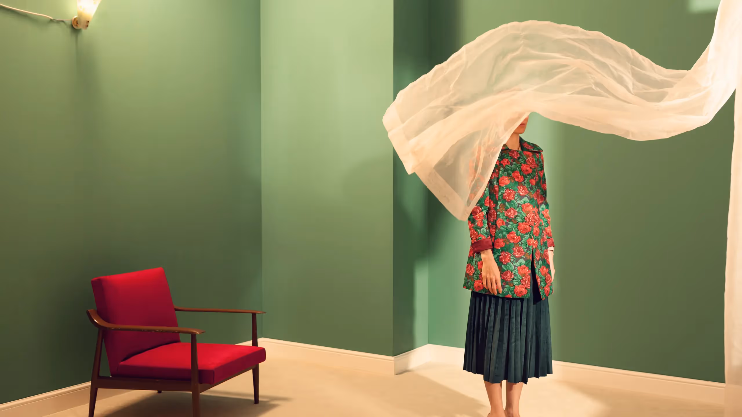 A woman stands in a green room with a red chair, her face concealed by a billowing curtain.
