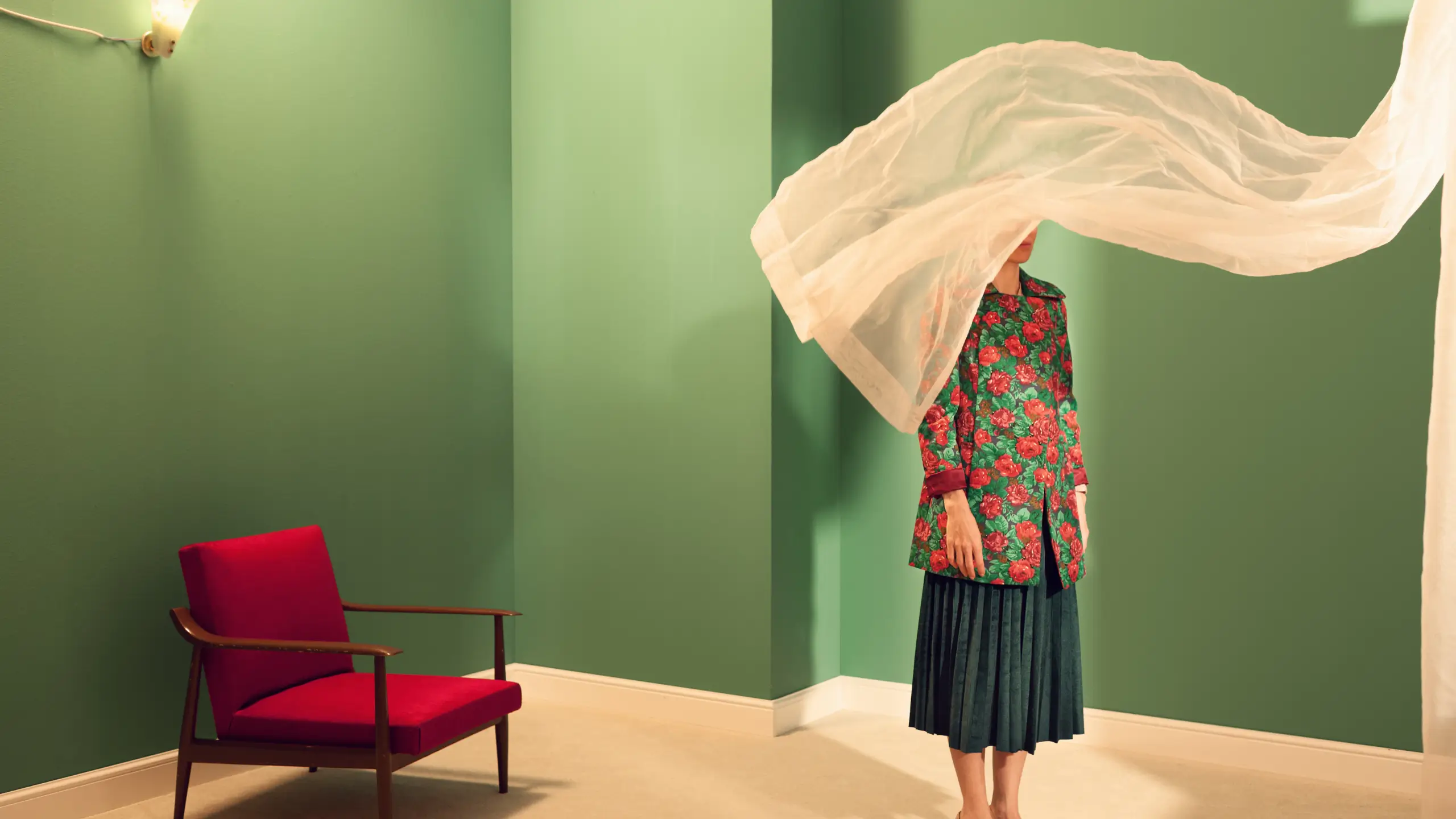 woman stands in a green room with a red chair, her face concealed by a curtain for a blog about literary fiction