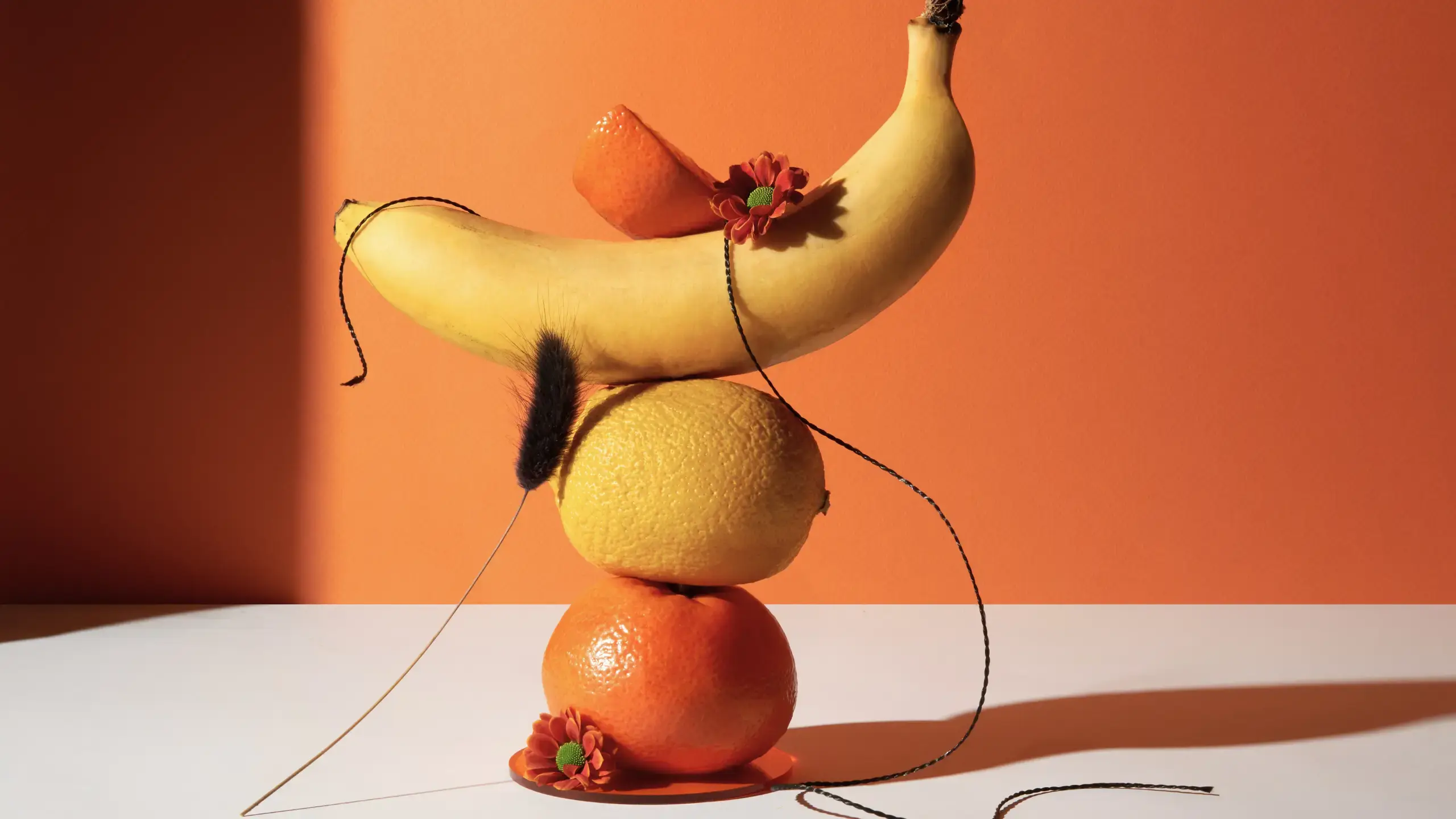A banana stacked on top of a lemon stacked on top of a small orange, with some red flowers winding around the fruits.