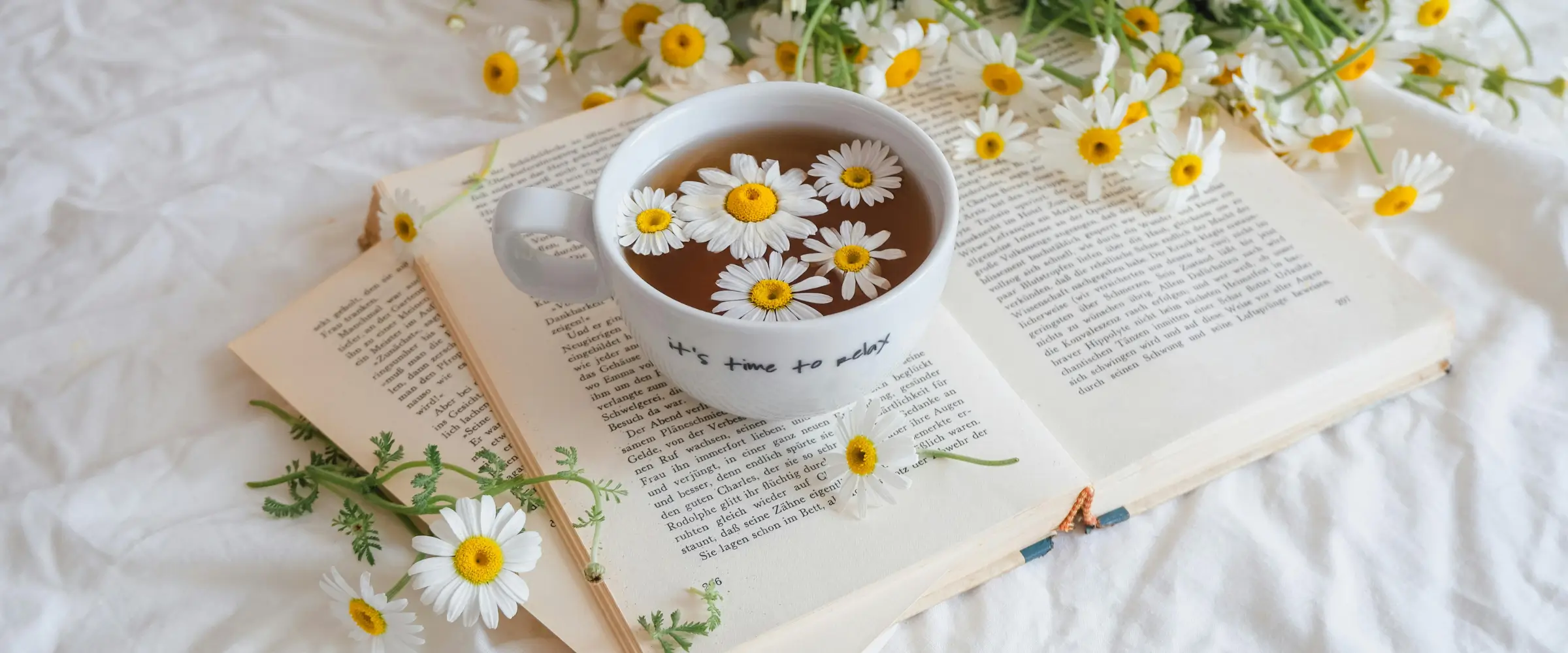 A book lies open on a muslin cloth, a cup of herbal tea resting on top of it along with many daisy flowers.