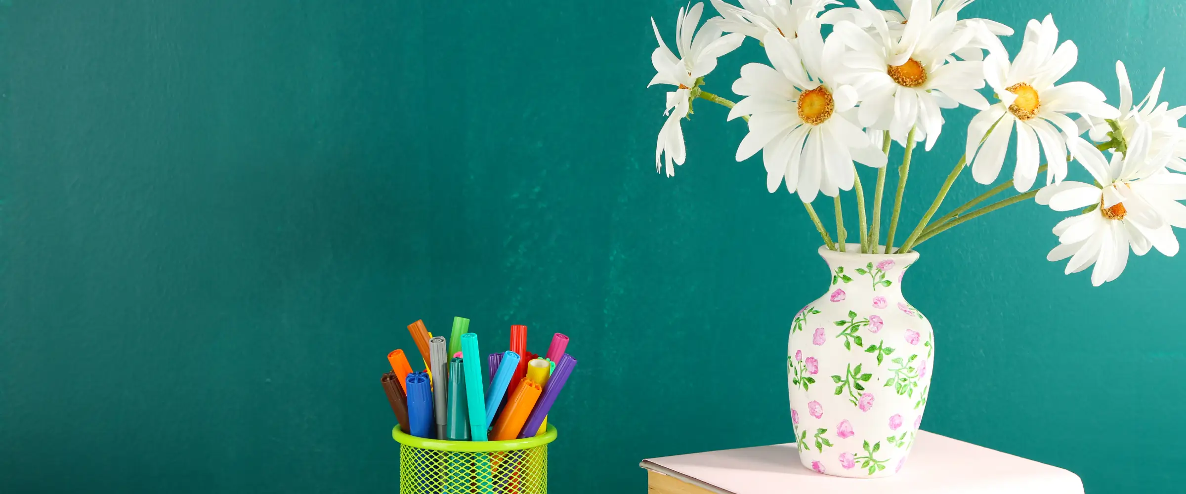 A pot of colored pens and a floral vase containing large white flowers sit against a turquoise backdrop.