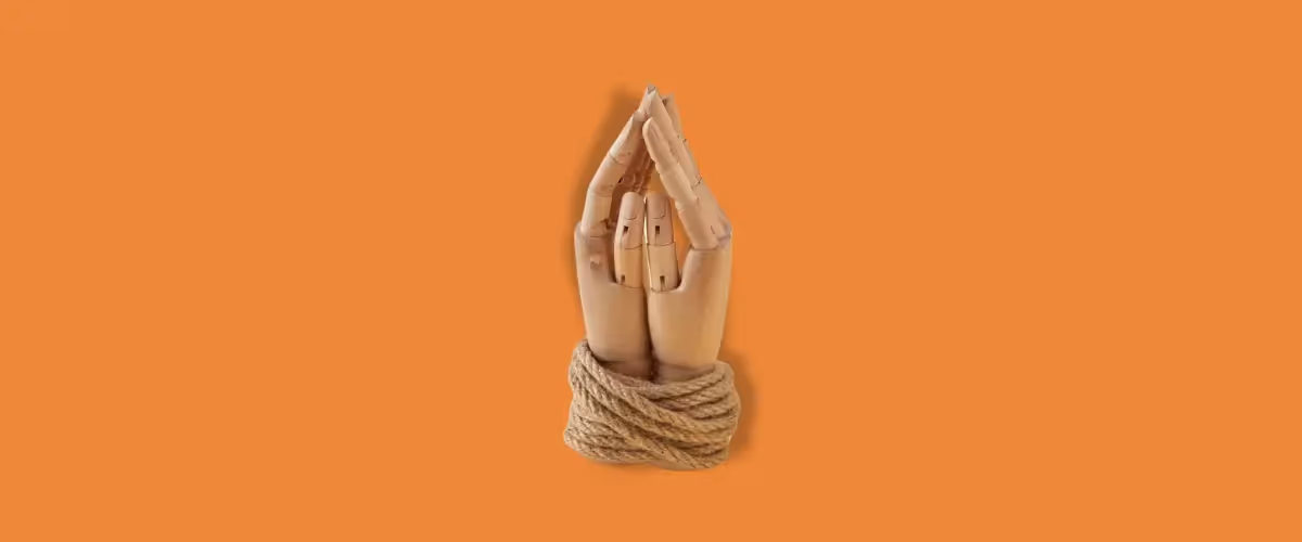 Wooden artist model hands clasped together and wrapped in rope against an orange background.