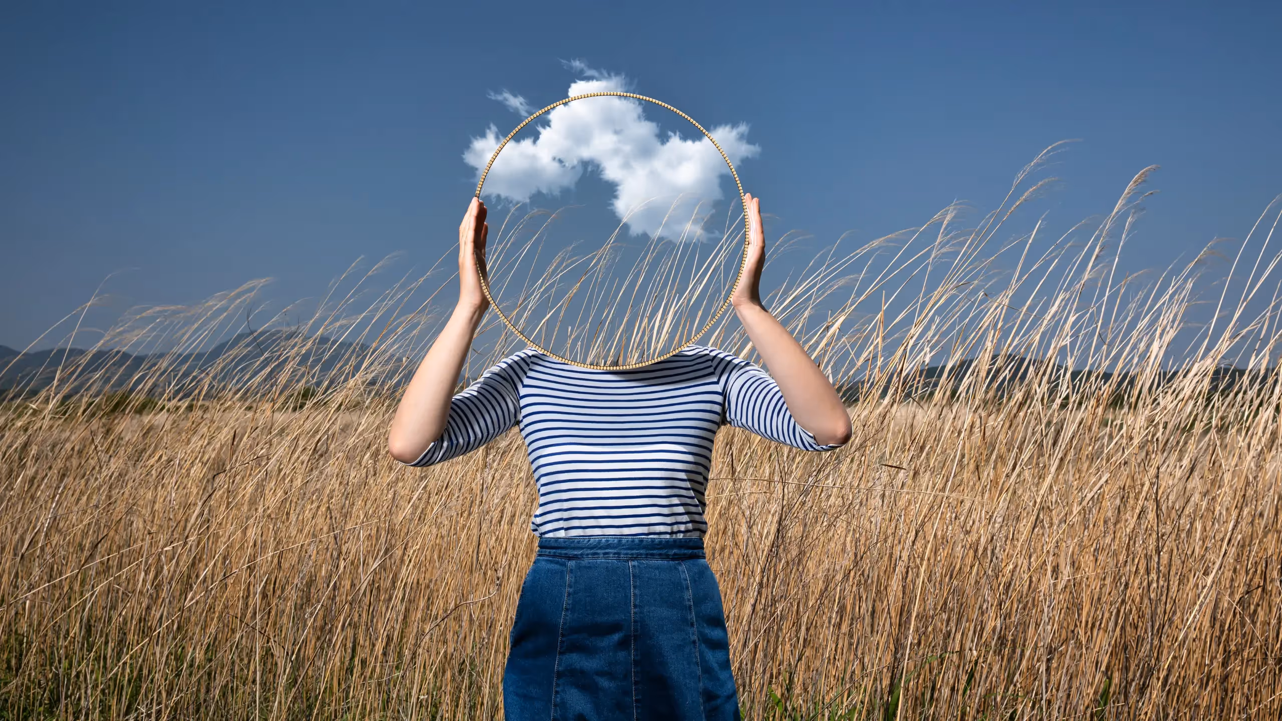 A woman holding a mirror in front of her face against a meadow background, reflecting a cloud-spotted blue sky.