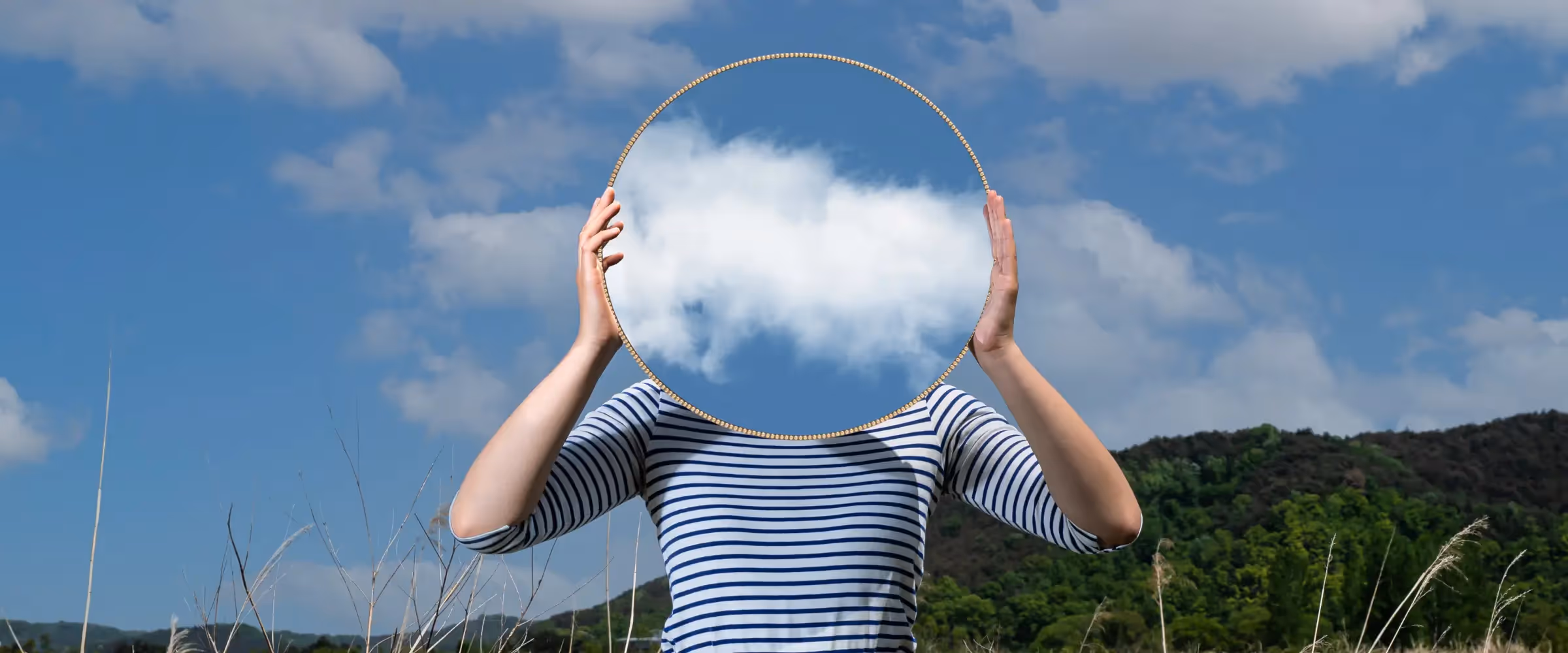 A person holding a round mirror in front of their face, hiding it by showing only a reflection of a cloud-spotted blue sky.