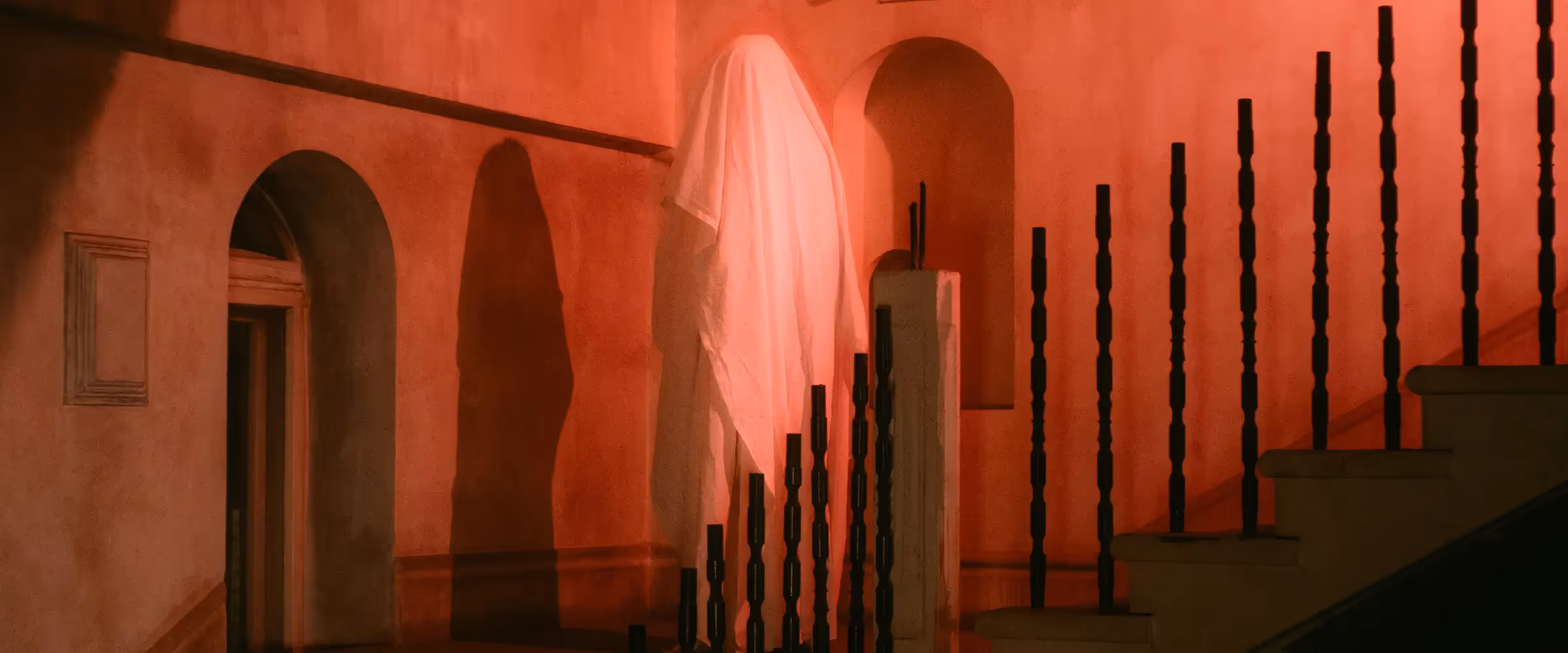 A figure cloaked in a white sheet stands on a staircase in red-tinted lighting.