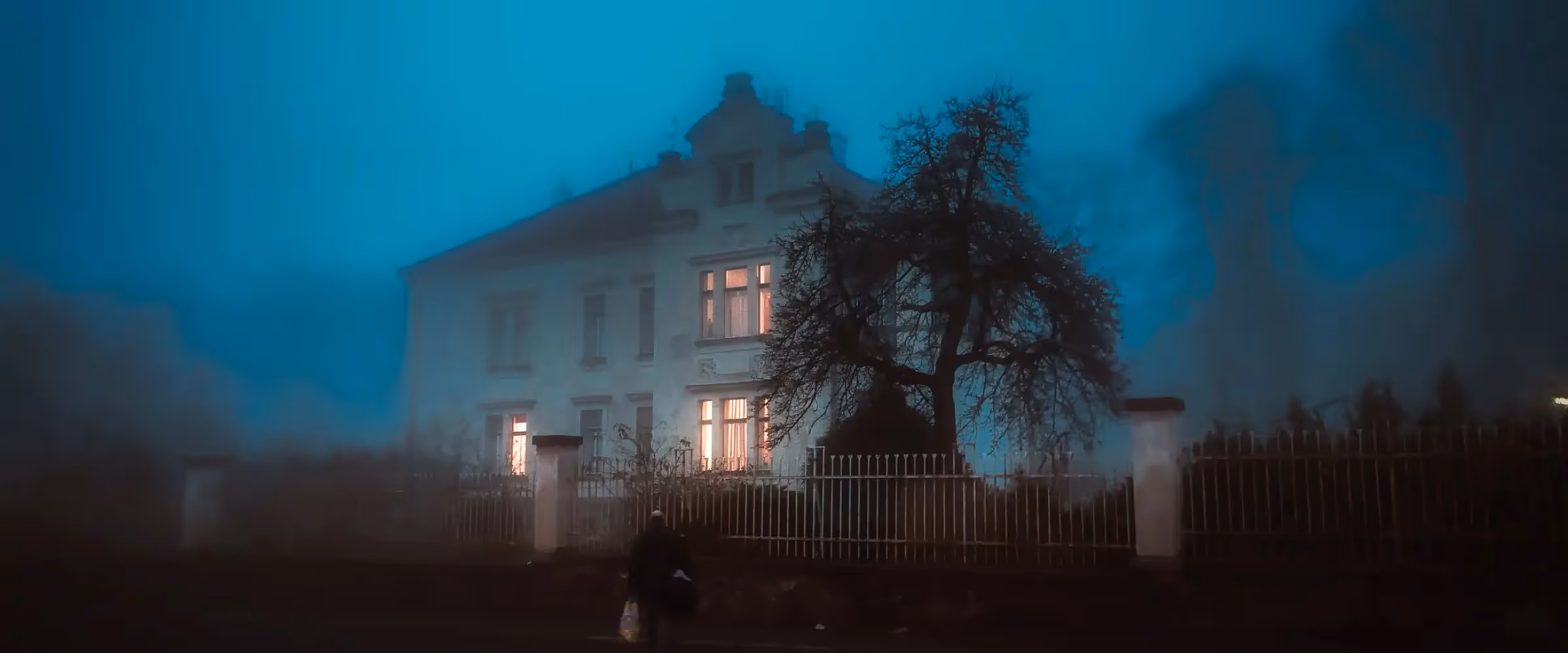 A house with lit windows behind an iron railing fence on a misty night.