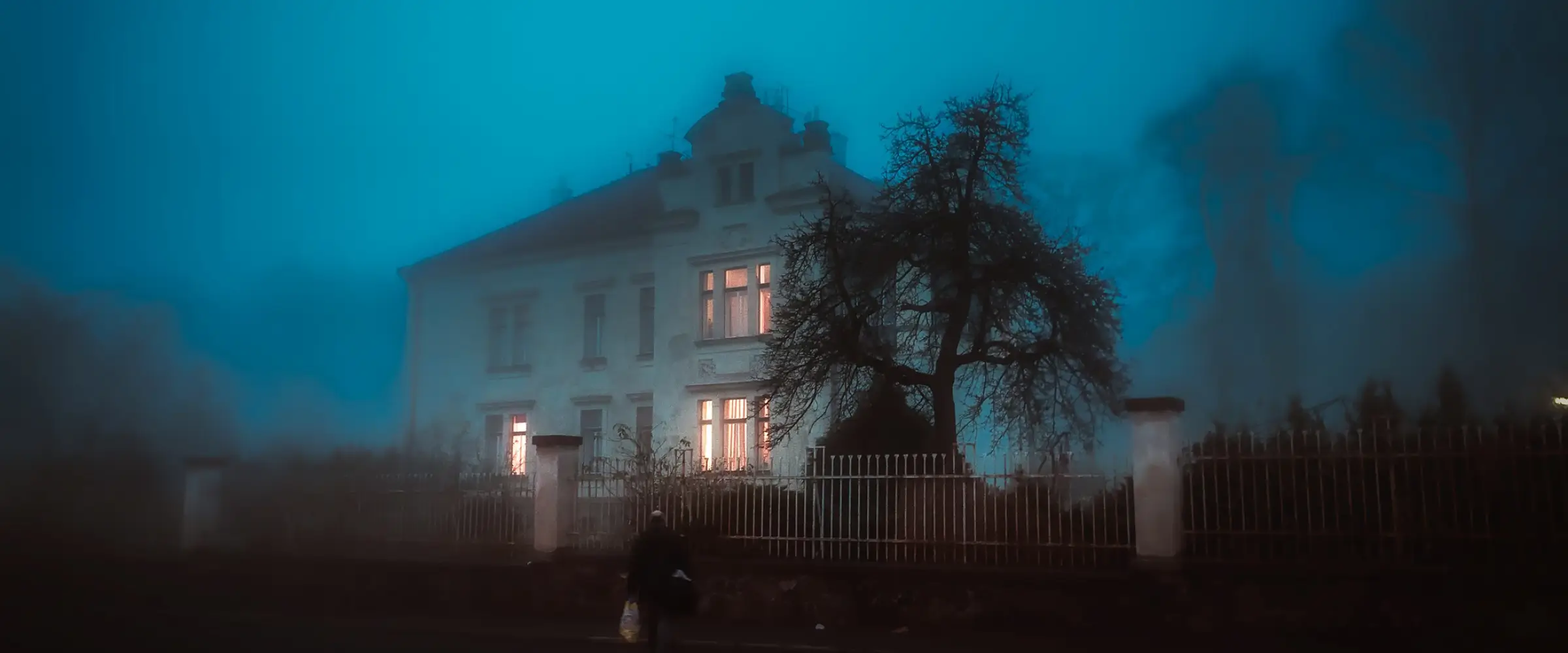 A house with lit windows behind an iron railing fence on a misty night.