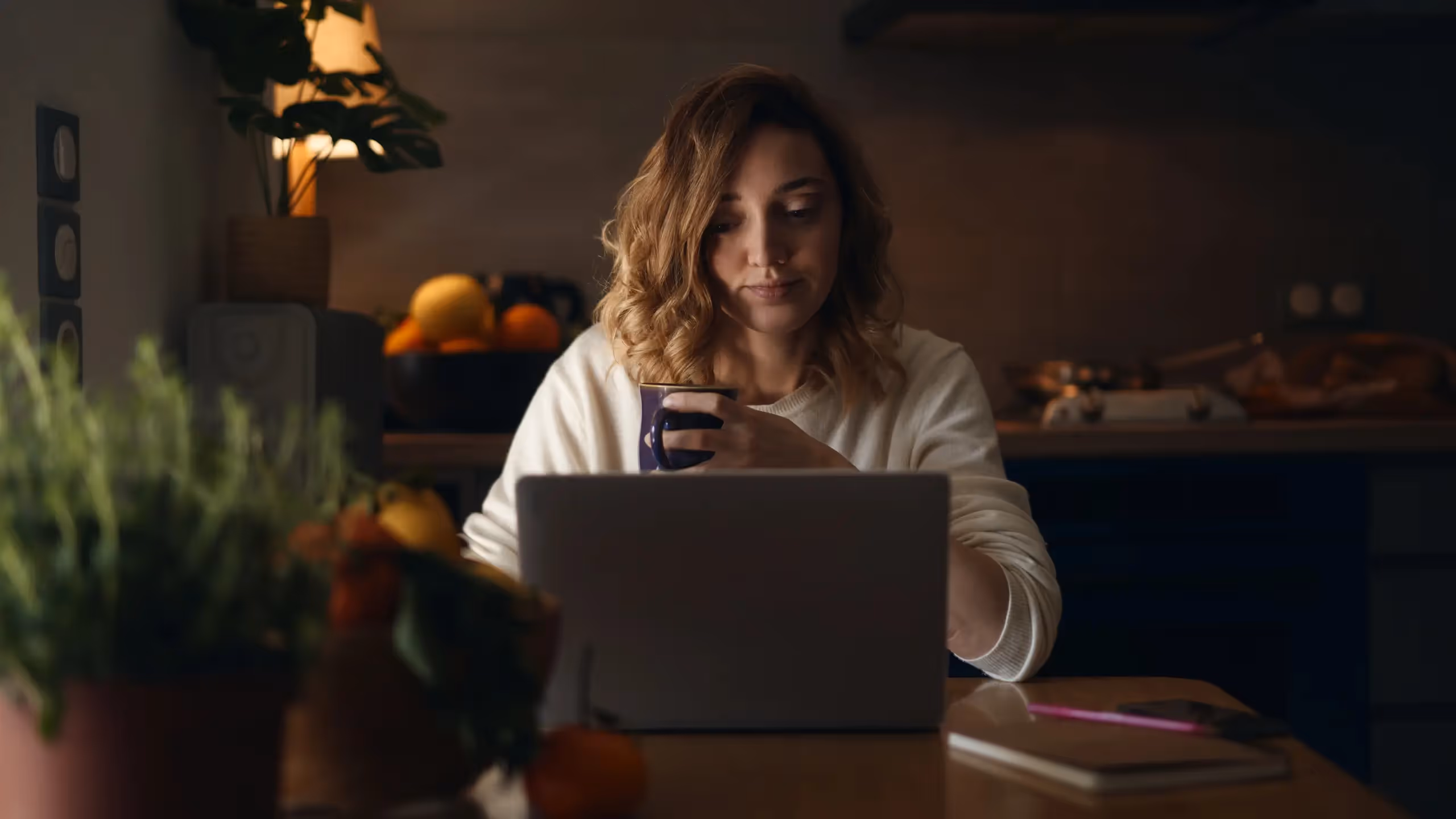 A woman works on a laptop in a dark kitchen while holding a mug.