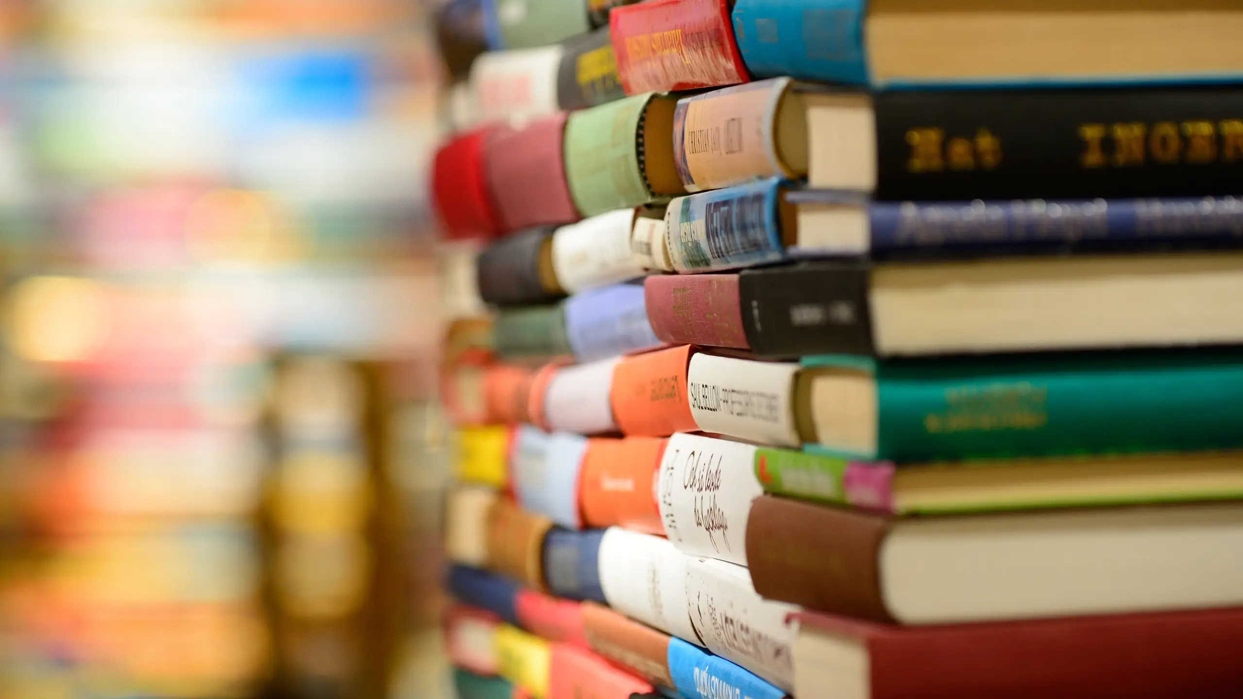 Spines of colorful books, stacked in a high pile with more in the background.