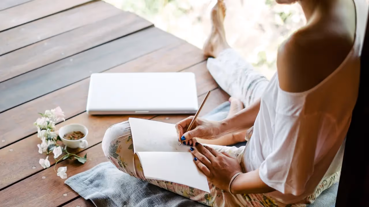 A woman sits on a wooden porch, writing in a notebook.