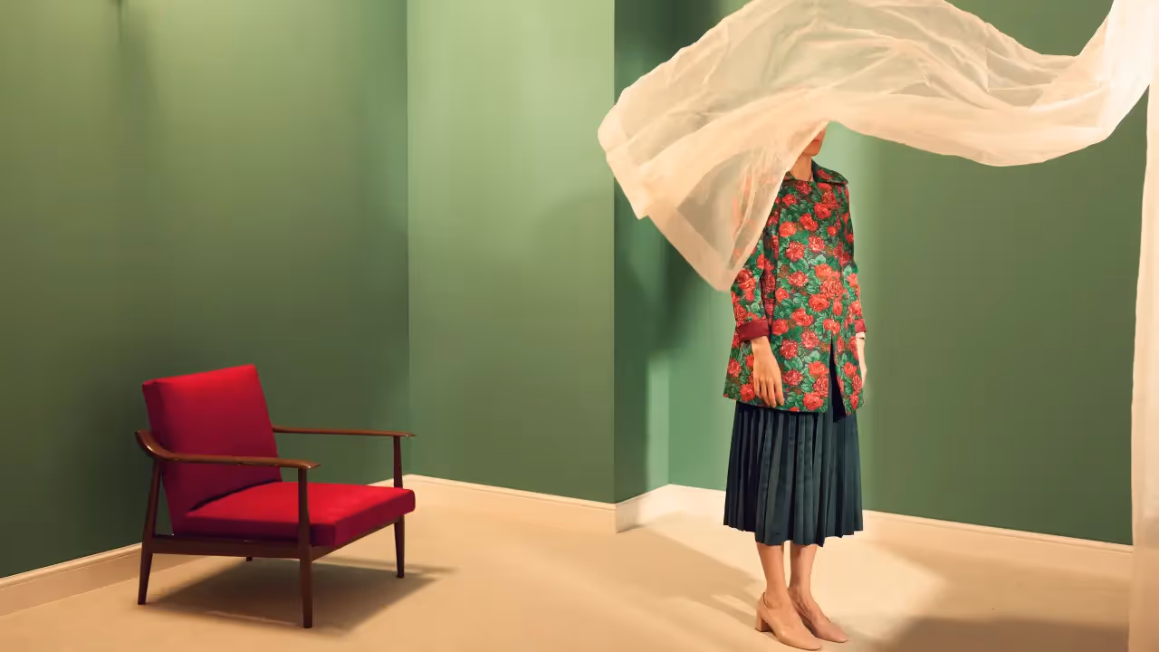 A woman wearing a floral coat stands in a room with a voile curtain blowing across her face.